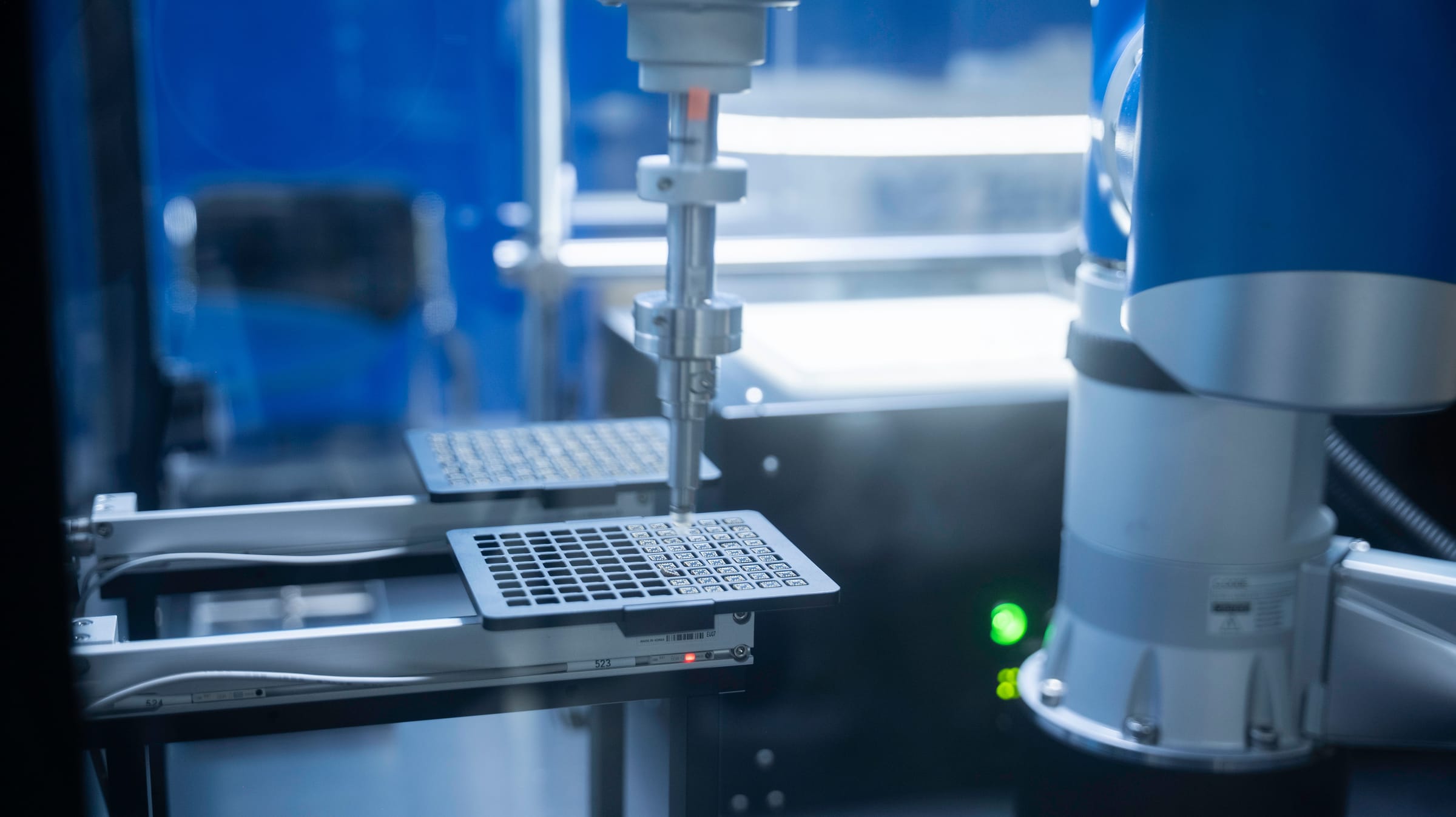 Automated laboratory arm lowering a thin probe over a sample plate inside a dim, blue-lit enclosure.