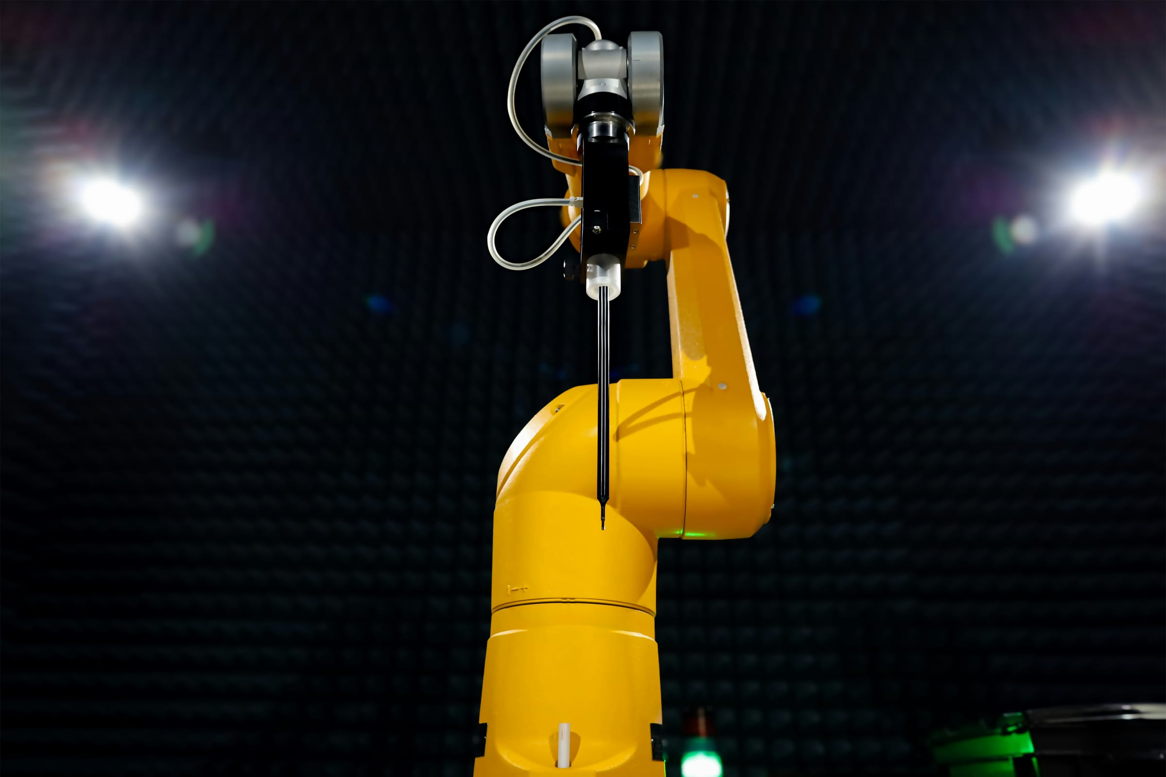A yellow six-axis robot arm holding a slender probe, lit by two lamps in a dark chamber lined with absorber foam.