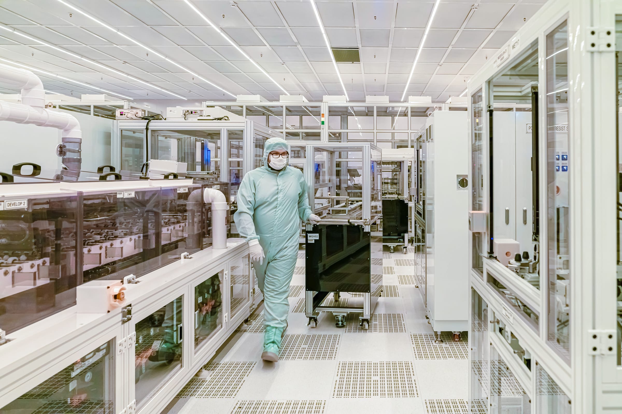 A technician in a full cleanroom suit, hood and mask walks along an aisle of enclosed manufacturing equipment in a brightly lit cleanroom.